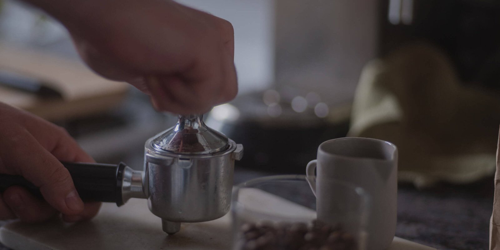 Una persona prensa posos de café con un tamper en un portafiltro junto a una taza de café blanca en la encimera de una cocina. Al fondo se ve un tarro de granos de café y una toalla.