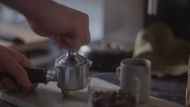 Una persona prensa posos de café con un tamper en un portafiltro junto a una taza de café blanca en la encimera de una cocina. Al fondo se ve un tarro de granos de café y una toalla.